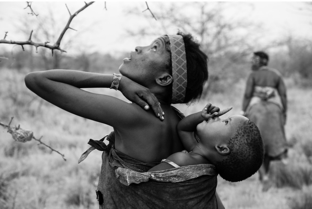 Black and white picture of a San mother carrying an infant on her back. She is pointing upwards, and the child is pointing, too.