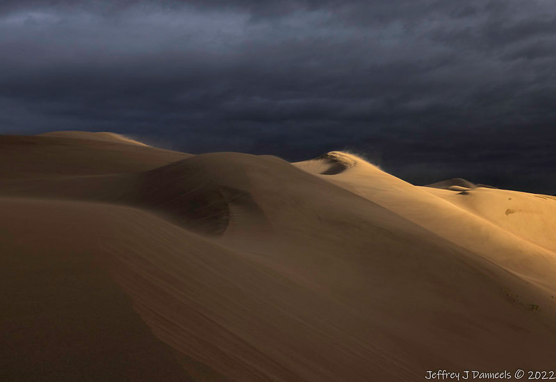 The tops of sand dunes with wind blowing over the tops of them, dark cloudy sky above.